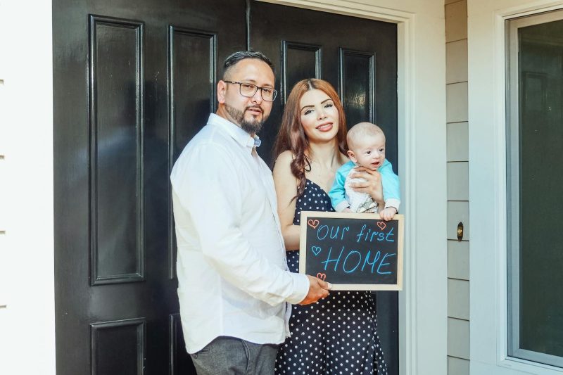Young family with baby holding a sign 'Our First Home' at doorstep, symbolizing new beginnings